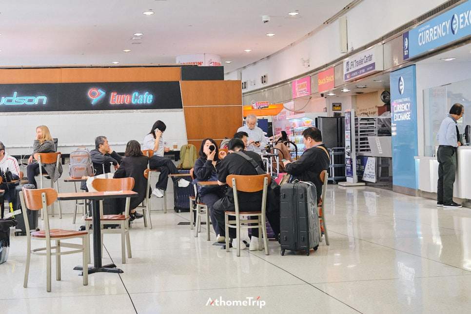 식당과 환전소가 있는 공항 내부 공간에 사람들이 앉아 있고 짐 가방이 놓여 있는 장면