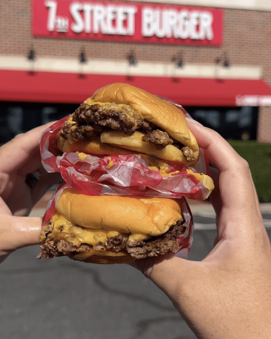 7th STREET BURGER 문구가 적힌 종이와 함께 두 개의 버거를 손으로 들고 있는 실외 장면