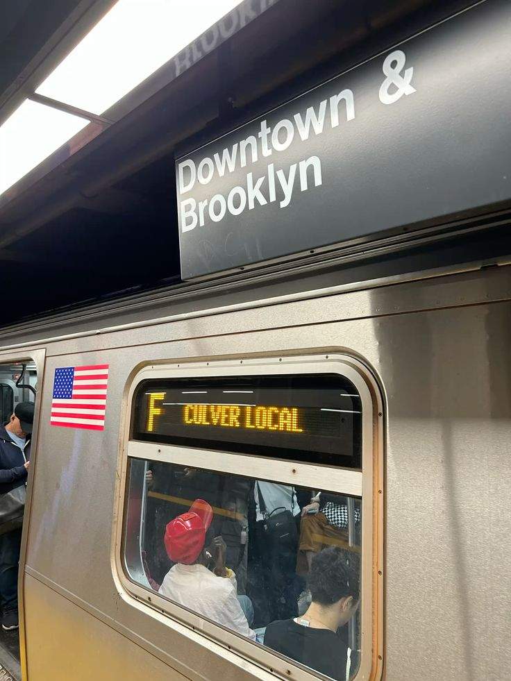열차 내부 창문 너머로 보이는 Downtown & Brooklyn 문구가 적힌 지하철 표시판과 미국 국기