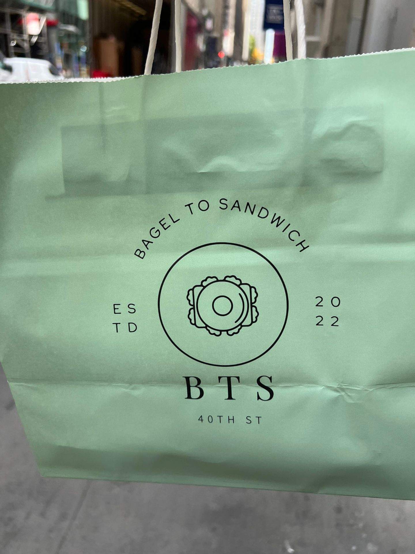 40TH ST 문구가 적힌 종이 봉투에 BTS와 BAGEL TO SANDWICH 로고가 보이는 실외 장면