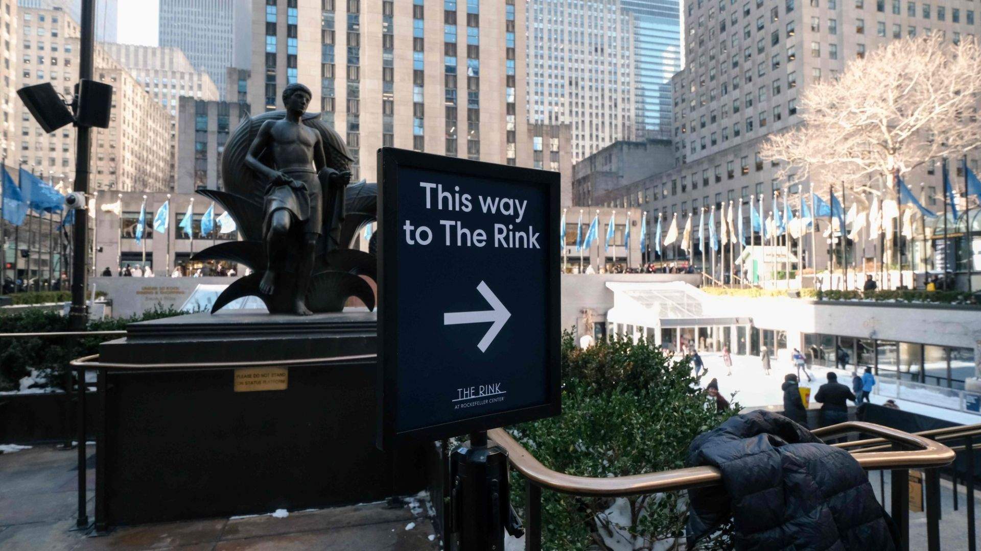 This way to The Rink라고 적힌 안내판과 광장의 조형물 및 건물이 보이는 실외 장면