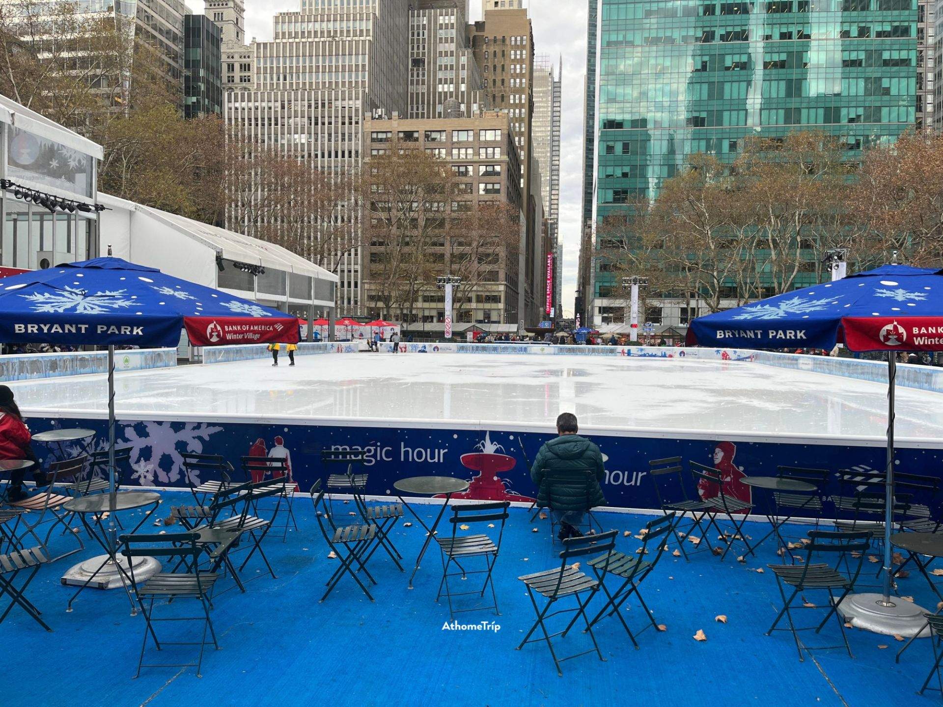 BRYANT PARK 빙판 주변에 배치된 테이블, 의자와 우산이 보이는 도시 공원