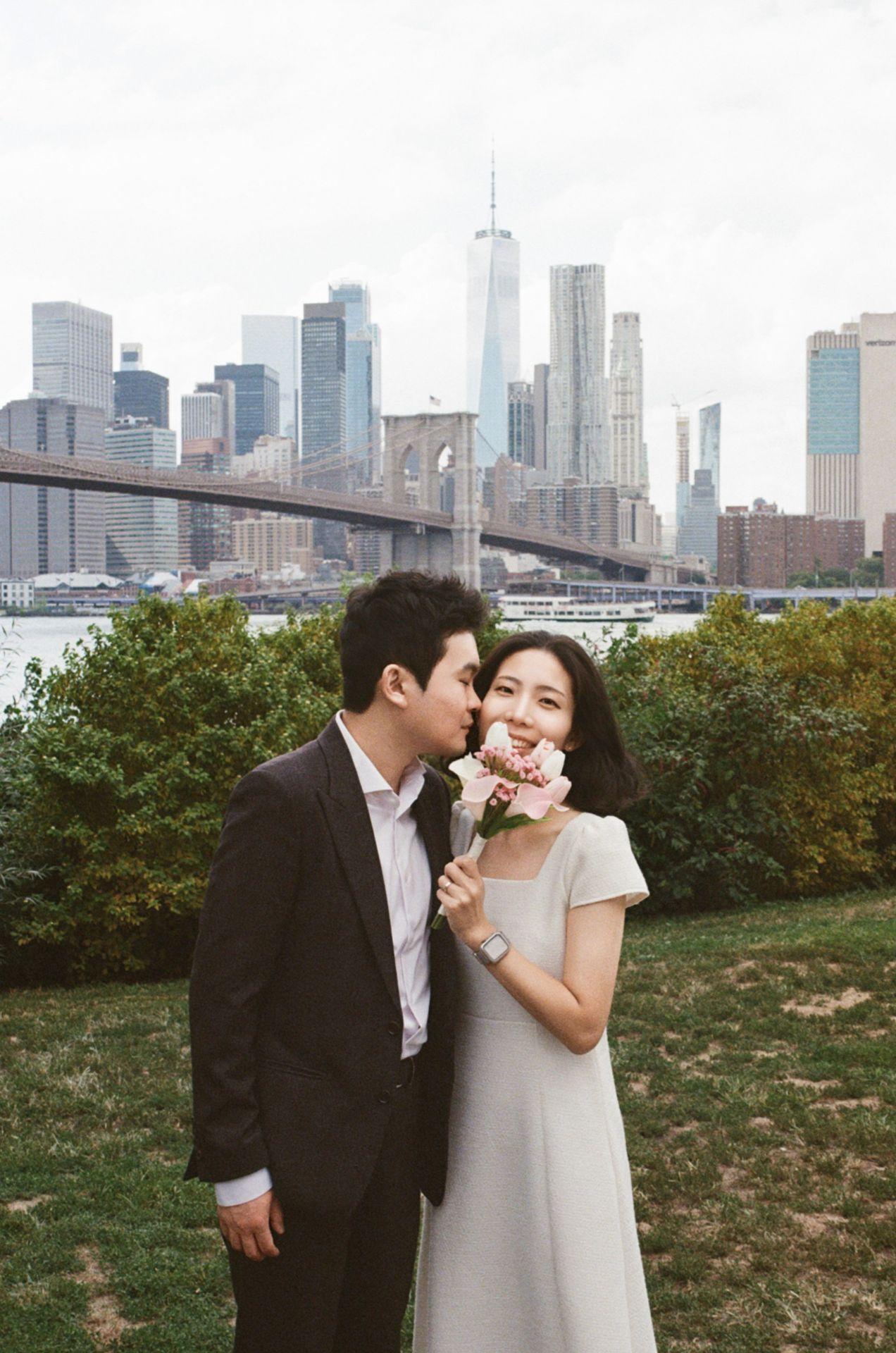 공원에서 꽃과 함께 도시의 건물 및 다리를 배경으로 서 있는 두 사람의 모습