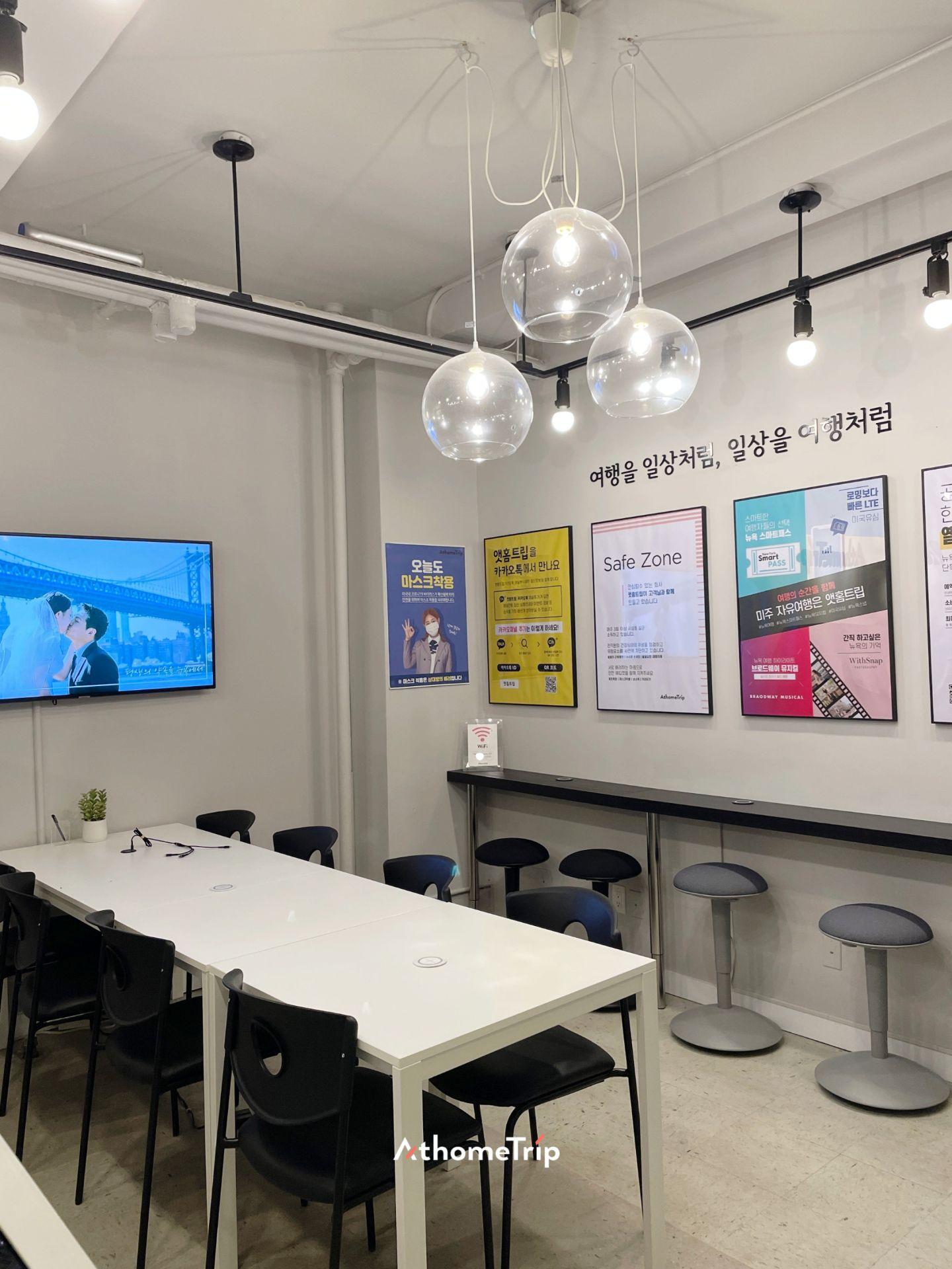 실내 공간에 흰색 테이블, 검은 의자, 벽면의 포스터와 전광판이 보입니다.
