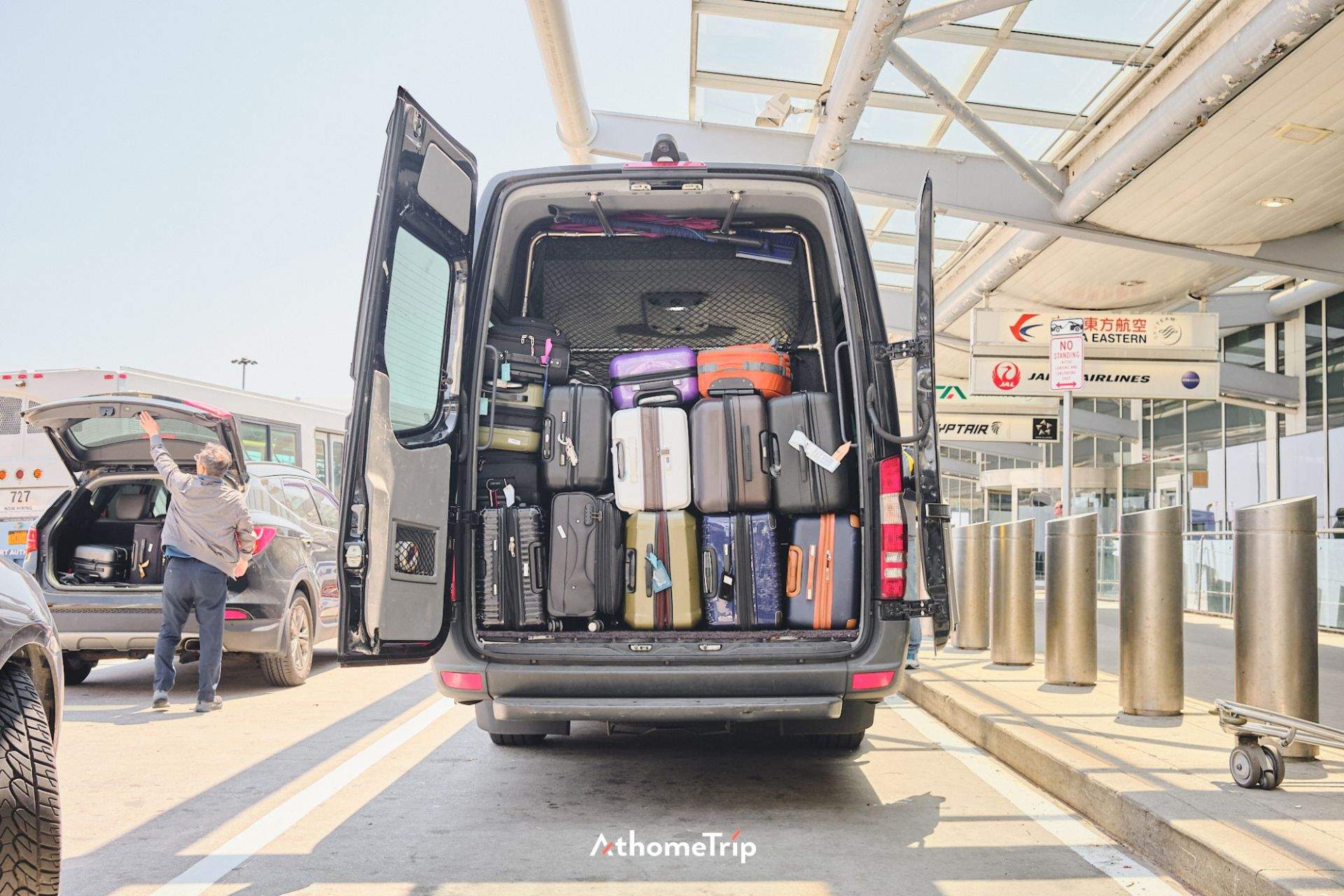 공항 주차장에 세워진 대형 차량의 후면 문이 열려 있으며, 내부에는 여러 개의 여행 가방이 밀집되어 있습니다. 주변에는 다른 차량과 공항 건물이 보입니다.